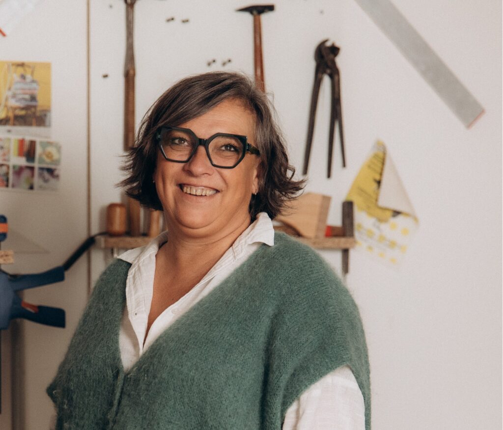 Portrait de Sabine Naudinat, fondatrice de l'atelier Au Fauteuil d'Inès tapissier artisanal dans son atelier, souriante devant ses outils et matériaux de rénovation de fauteuils. Portant un pull vert amande sans manche avec une chemise blanche et des lunettes vertes