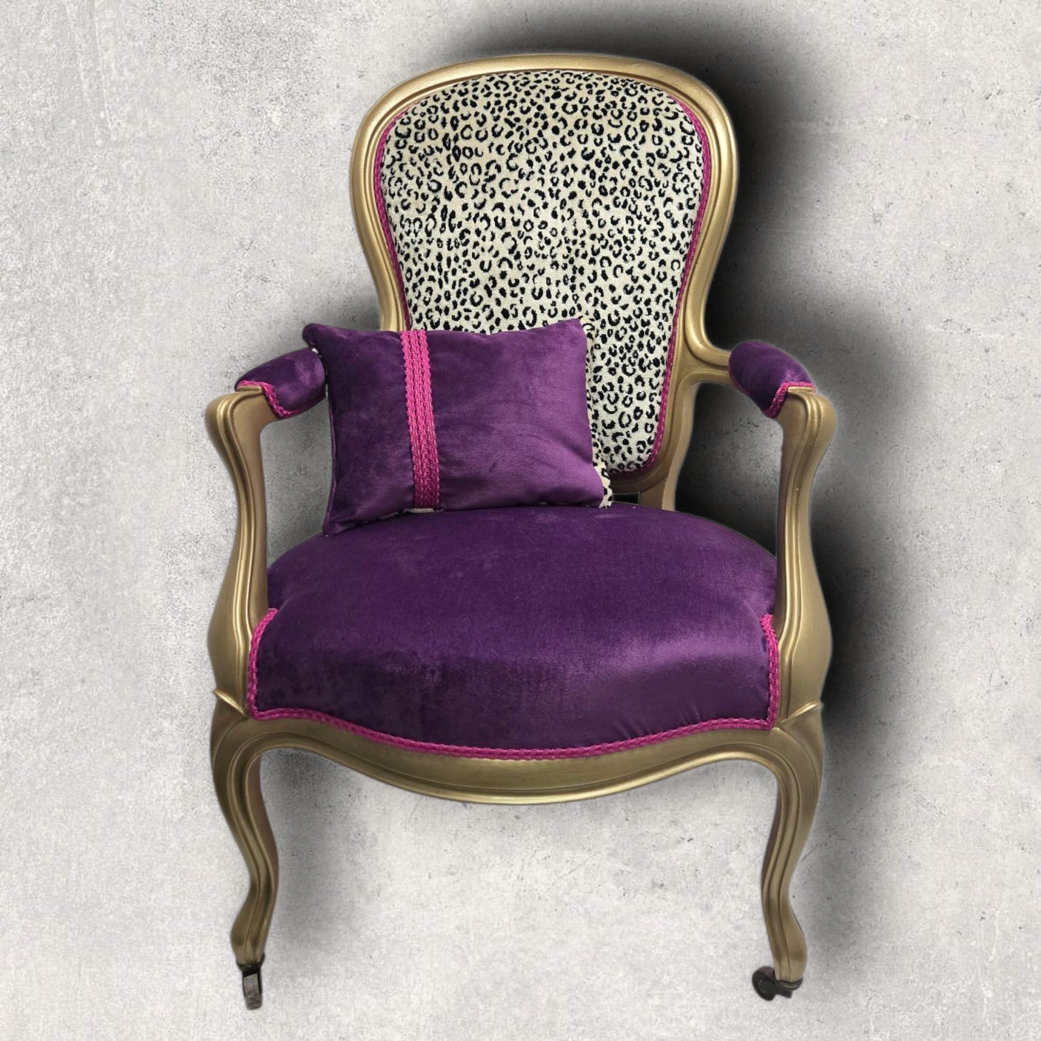 Fauteuil de style Louis XV ou Bergère, restauré avec un style contemporain et audacieux, sur un fond texturé gris clair. Le cadre en bois est peint ou recouvert d'une finition dorée ou laiton. L'assise est recouverte d'un velours violet profond, bordé d'un galon décoratif rose fuchsia. Le dossier est tapissé d'un tissu à motif imprimé léopard noir sur fond blanc/crème. Un petit coussin carré en velours violet, avec une bande décorative rose, est posé sur l'assise.
