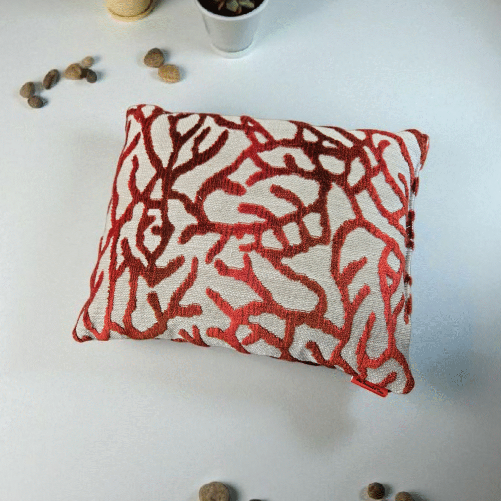 coussin décoratif carré. Le tissu est blanc cassé avec un motif en relief qui ressemble à des branches de corail ramifiées, de couleur rouge et bordeaux, sur un fond noir uni. Le motif est texturé, en velours contrastant avec le fond tissé. le fond est blanc avec des cactus en pot et quelques galets posés en décoration