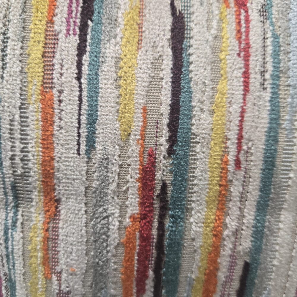 détail du coussin décoratif carré, dont la housse présente un motif à rayures verticales et irrégulières sur un fond beige-gris clair. Les rayures sont de couleurs vives, incluant le rouge, le jaune, le bleu sarcelle, le violet et l'orange. La texture du tissu est velours, ressemblant à un tissage chenille ou un jacquard texturé.