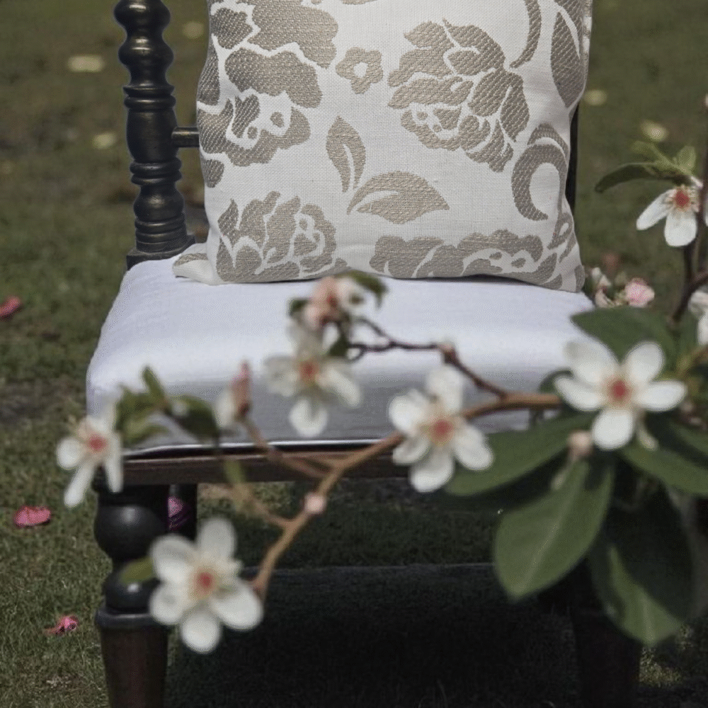 Coussin décoratif beige et blanc avec un motif floral en jacquard posé sur l'assise blanche d'une chaise en bois sombre et tournée, placée sur une pelouse verte. Un rameau fleuri avec des fleurs blanches est au premier plan, flou.
