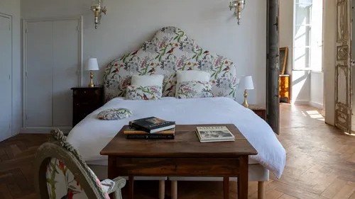 Chambre à coucher lumineuse avec une grande tête de lit capitonnée à motif floral d'oiseaux et de fleurs, des draps blancs, et une table basse en bois ancien. Sol en parquet chevrons. Style classique chic.